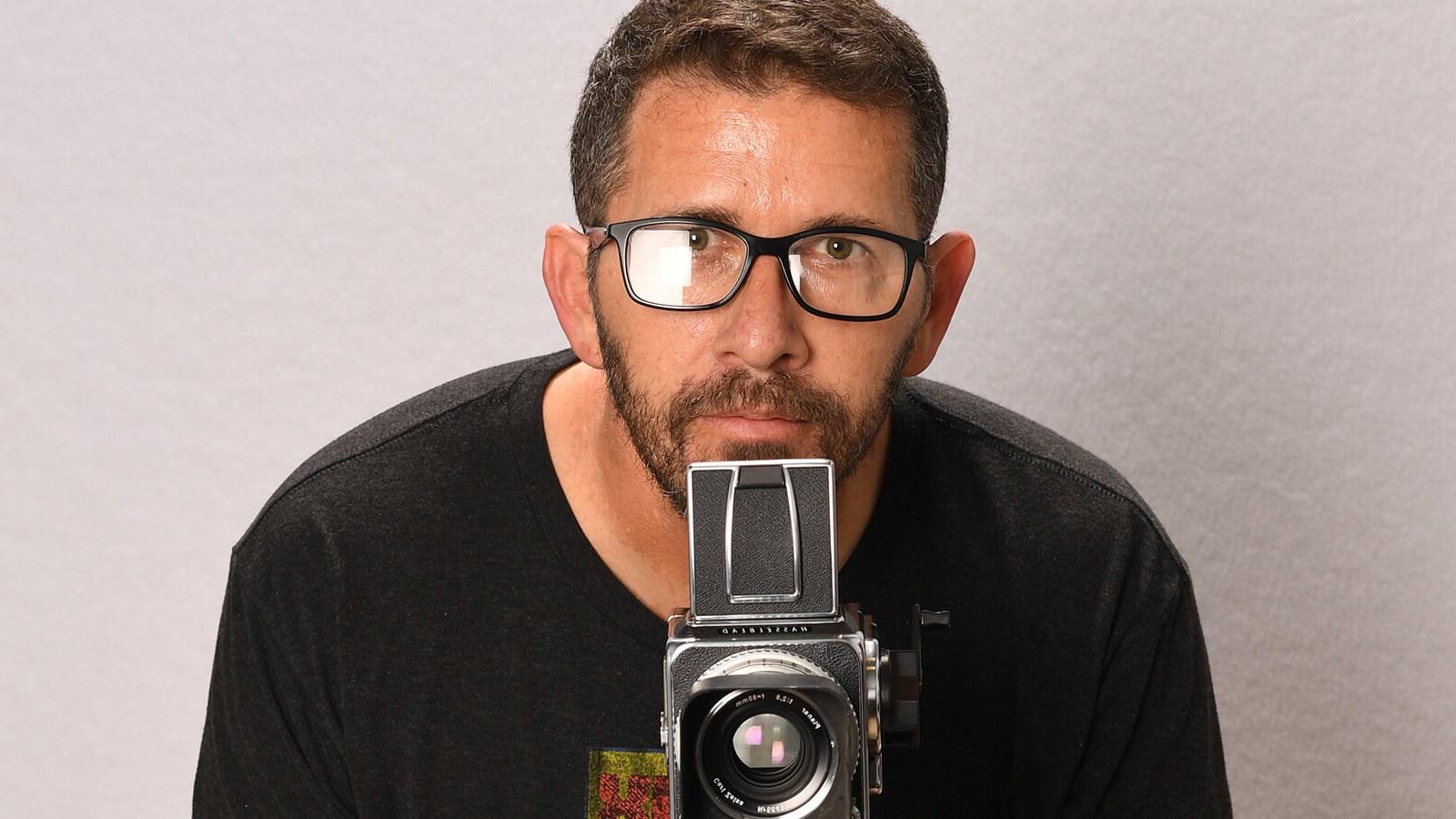 Sebastian Krys crouches behind a video camera. He is wearing black-rimmed eyeglasses and a black t-shirt with a logo on it.