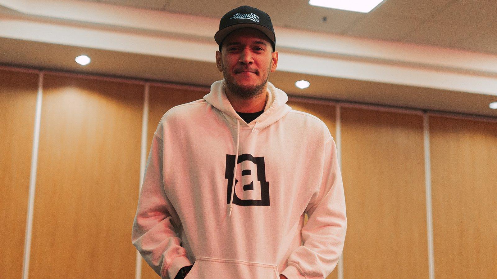 A man standing against a wood paneled wall in a white hooded sweatshirt featuring the Bandits logo and a black Bandits Gaming snapback cap.