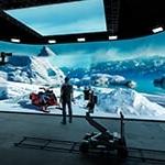 一个穿着牛仔裤、运动鞋和格子衬衫的男人站在制作工作室里. 他面对着一个巨大的LED屏幕，屏幕上显示着冰封的山脉和雪地摩托.