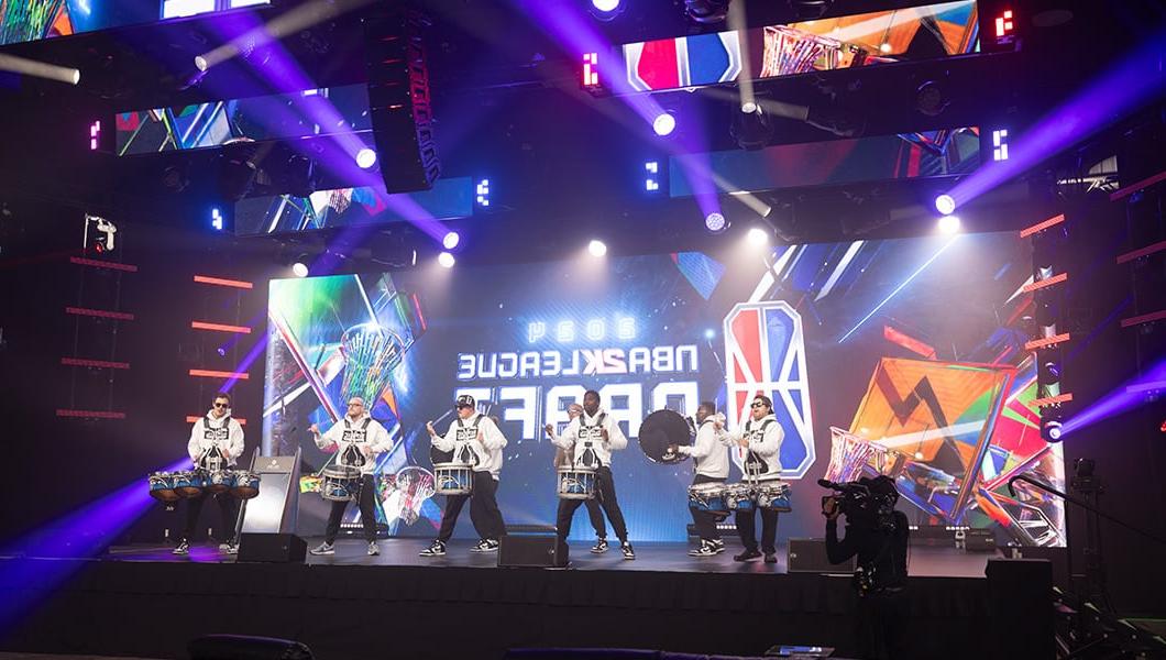 A drumline is performing on a stage in front of a large LED screen that reads “NBA 2K League Draft.”